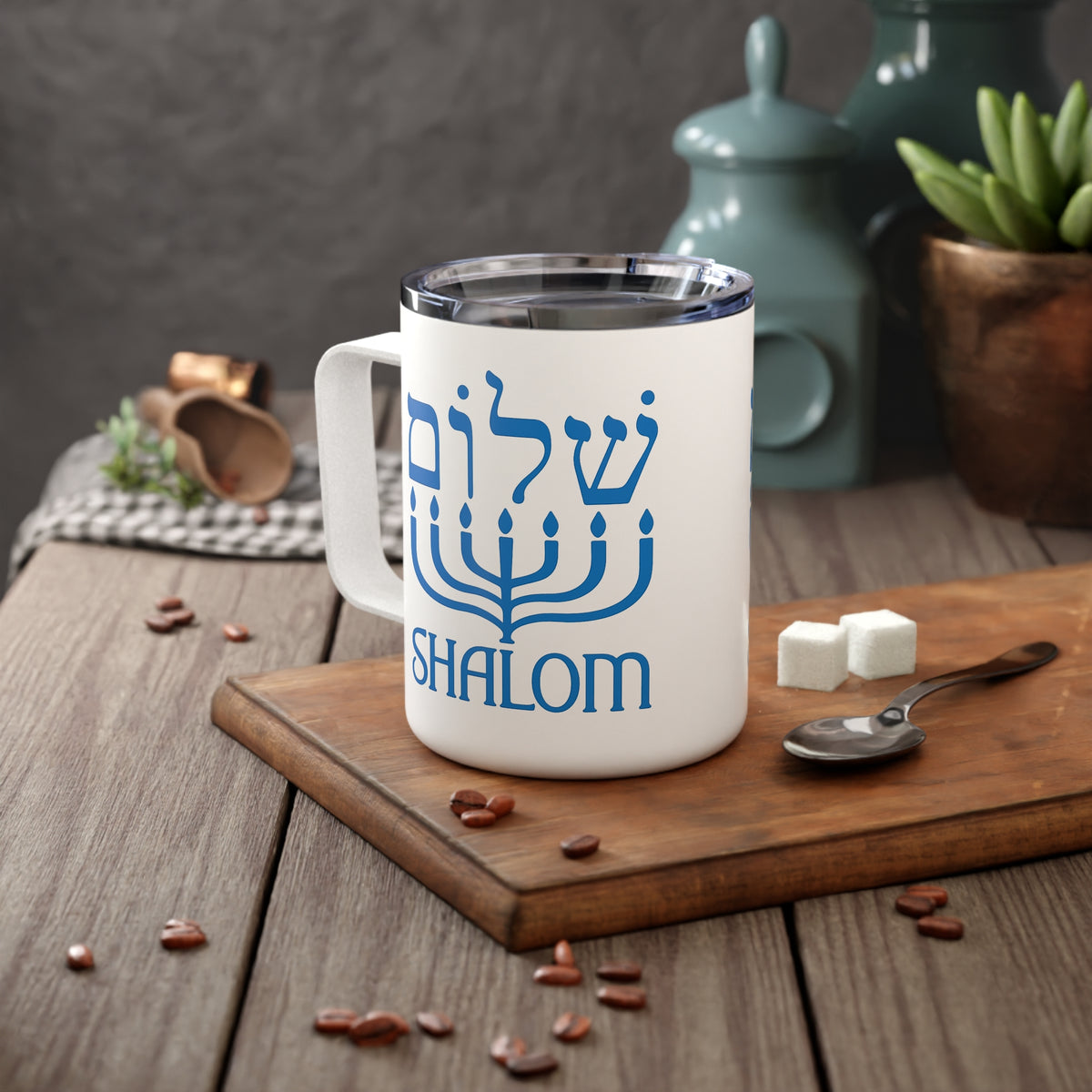 A white stainless steel  mug with a blue menorah graphic and the word 'SHALOM' on the front, placed on a wooden cutting board on a rustic table with coffee beans, sugar cubes, a spoon, and blurred teal jars in the background.