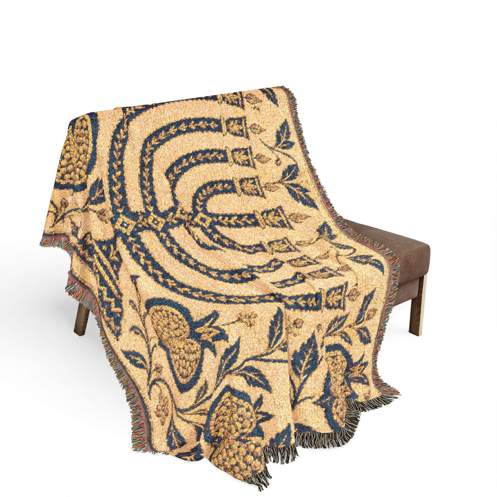 A woven throw blanket with a fringed edge draped over a brown upholstered chair against a plain white background. The blanket features a large-scale pattern in navy blue and tan with pomegranate motifs and curved geometric bands.