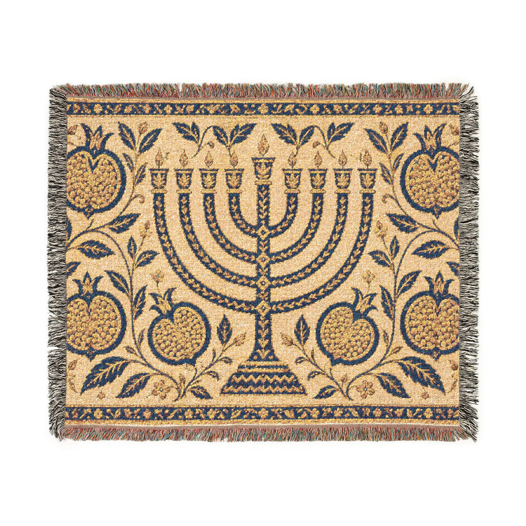 A rectangular woven textile with fringe on all sides, featuring a central menorah surrounded by pomegranate motifs and floral vines on a warm beige background. The design is primarily navy blue with tan and muted red accents in the fringe and border details.