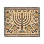 A rectangular woven textile with fringe on all sides, featuring a central menorah surrounded by pomegranate motifs and floral vines on a warm beige background. The design is primarily navy blue with tan and muted red accents in the fringe and border details.