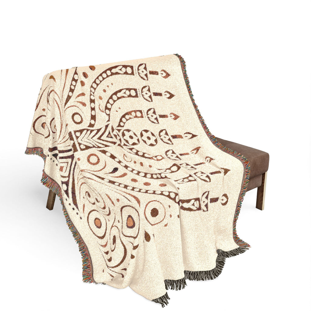 A woven throw blanket with a cream base and brown geometric tribal-style patterns, draped over a brown upholstered bench against a white background.