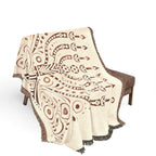 A woven throw blanket with a cream base and brown geometric tribal-style patterns, draped over a brown upholstered bench against a white background.