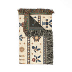 A folded woven throw blanket with fringe, featuring a cream base with blue, brown, and green geometric and floral patterns, shown on a plain white background.