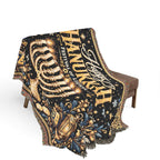 A woven throw blanket draped over a brown upholstered bench, featuring a black background with gold and blue festive illustrations and large decorative text, photographed on a clean white studio background.