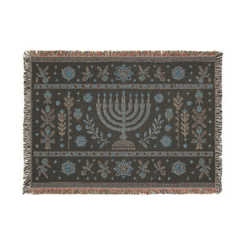 A rectangular woven textile with fringe on all sides, featuring a central menorah motif and symmetrical floral and botanical patterns in muted blue, beige, and brown on a dark charcoal background.