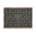 A rectangular woven textile with fringe on all sides, featuring a central menorah motif and symmetrical floral and botanical patterns in muted blue, beige, and brown on a dark charcoal background.