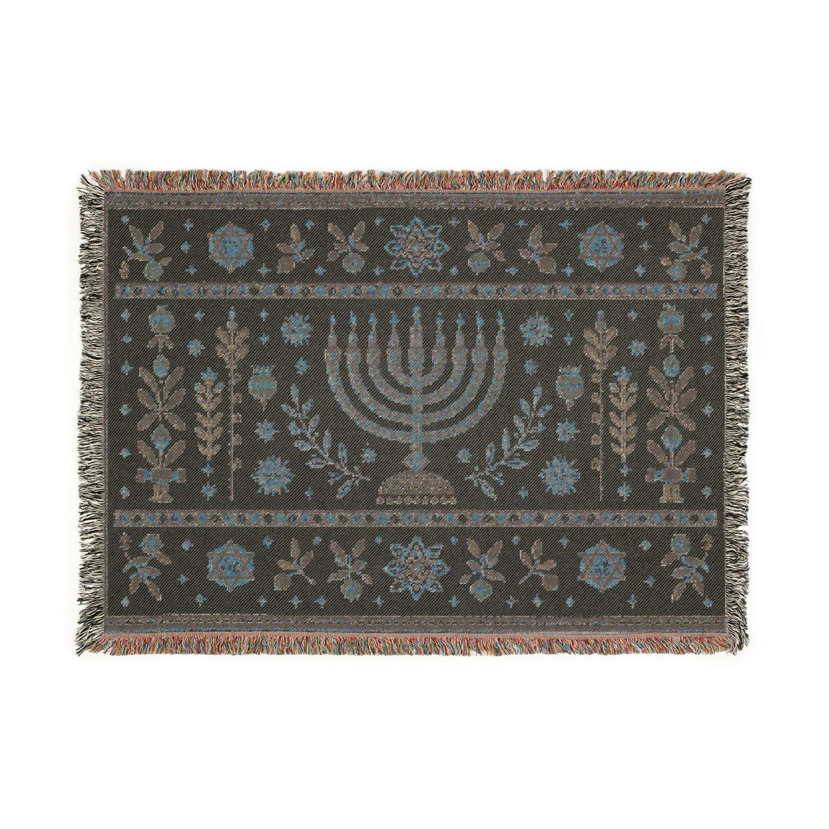 A rectangular woven textile with fringe on all sides, featuring a central menorah motif and symmetrical floral and botanical patterns in muted blue, beige, and brown on a dark charcoal background.
