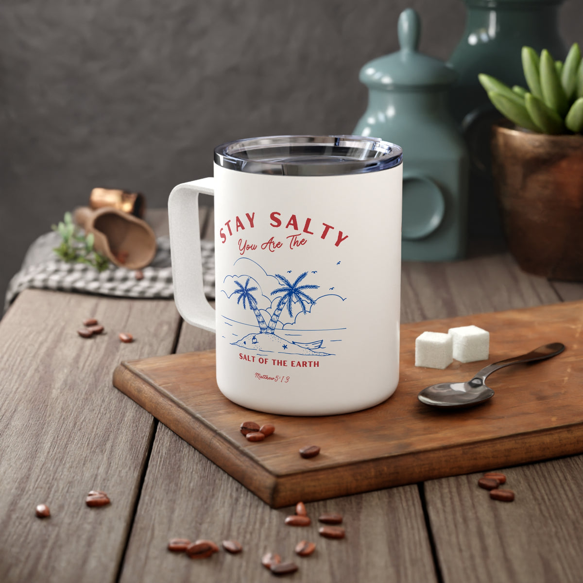A white insulated mug with a clear lid sits on a wooden board on a rustic table. The mug has a blue tropical palm tree illustration and red text reading “STAY SALTY You Are The SALT OF THE EARTH.