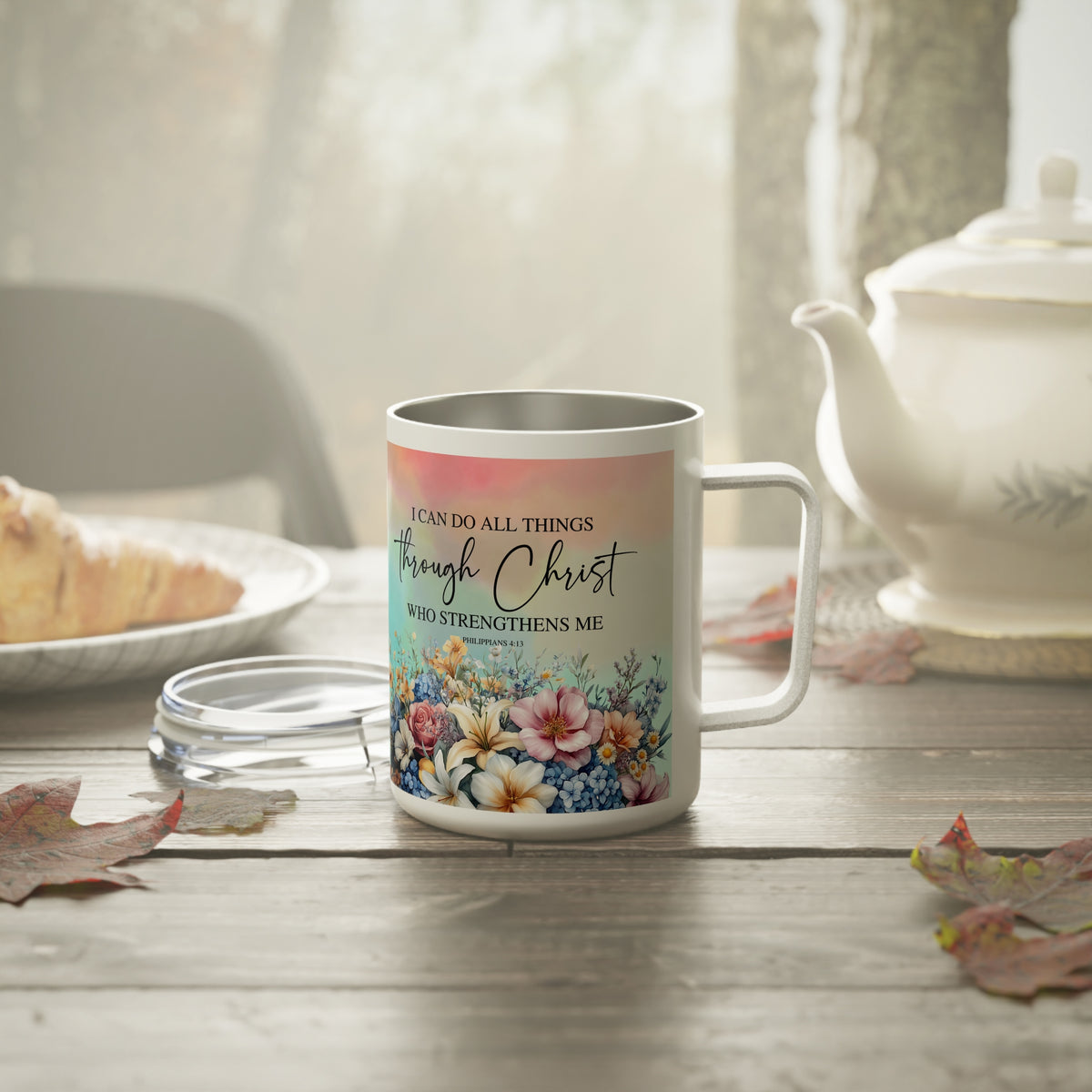 A stainless steel mug with a floral design and inspirational scripture text sits on a rustic wooden table next to a teapot and plate, with soft natural light filtering through a window in the background.