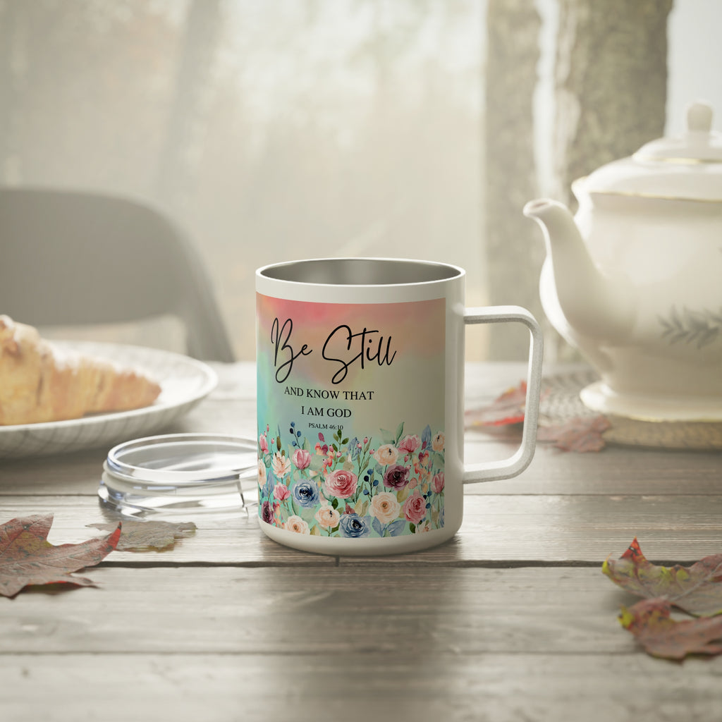 A stainless steel  mug with a floral design and the text 'Be Still AND KNOW THAT I AM GOD' sits on a wooden table in a softly lit kitchen or tea-room setting, with a teapot, plate, and leaves around it.