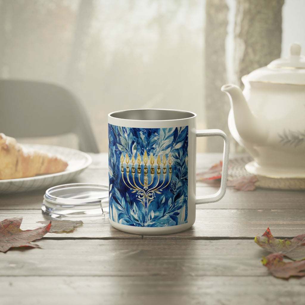 A white stainless steel mug with a blue and white floral pattern and a gold menorah design, set on a wooden table with a teapot and plate in a softly lit kitchen setting.