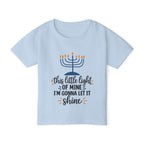 A light blue short-sleeve kids' t-shirt with a Hanukkah graphic on the front. The graphic features a navy blue menorah with orange flames and the text 'this little light OF MINE I'M GONNA LET IT shine' in navy and orange lettering.