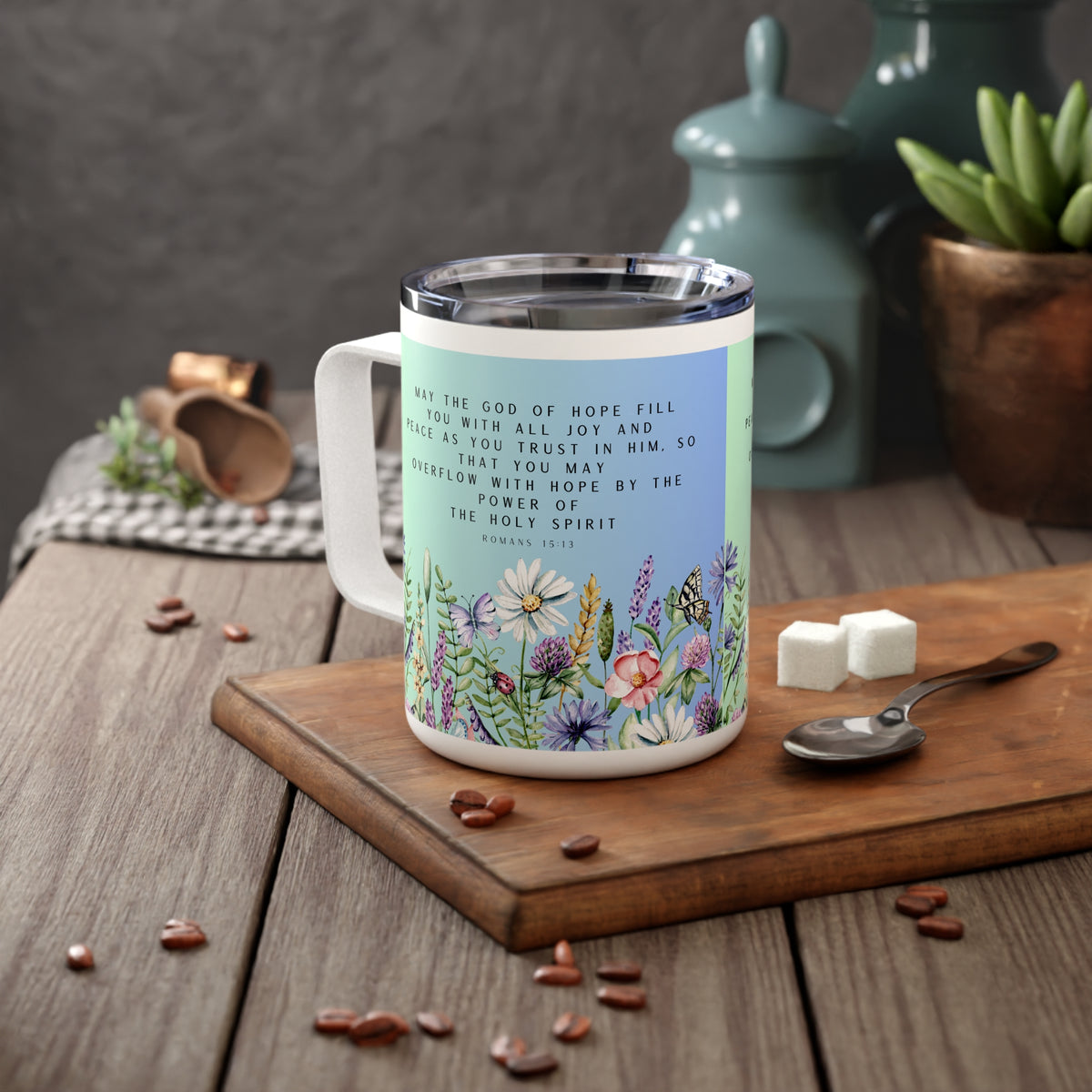 A white stainless steel coffee mug with a clear lid sits on a wooden cutting board on a rustic table. The mug features a light blue upper section with black Bible verse text and a lower section decorated with colorful wildflowers.