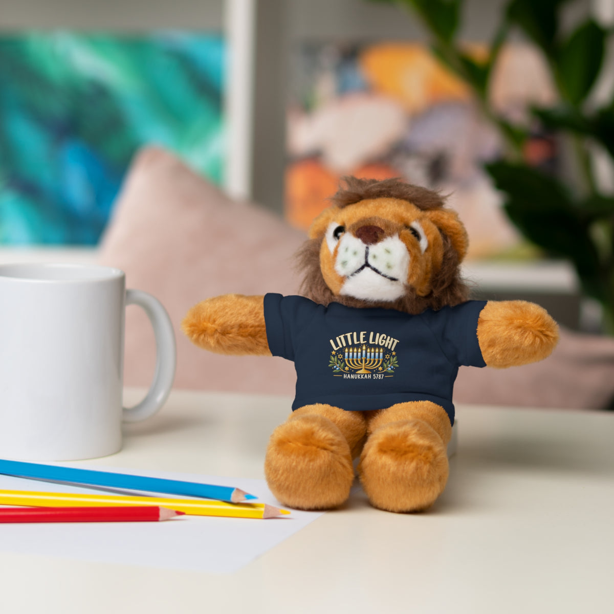 Plush lion toy wearing a t-shirt with a design, sitting on a table with a mug and pencils.