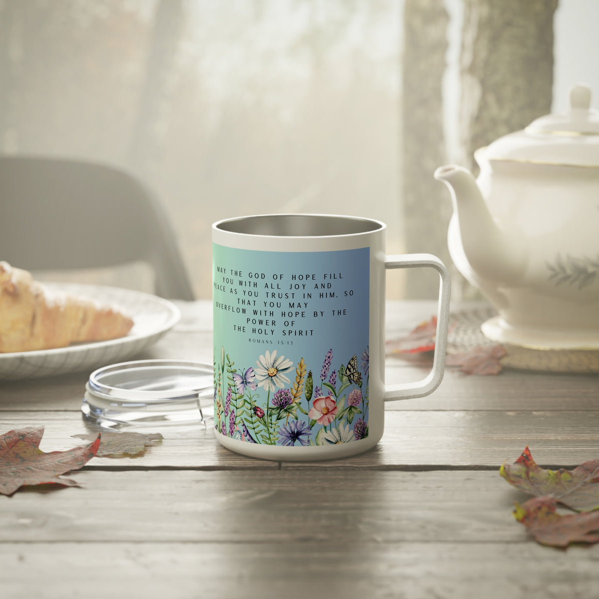 A white stainless steel coffee mug with a light blue front panel printed with a Bible verse and colorful wildflowers, set on a rustic wooden table with a teapot and plate in the background.