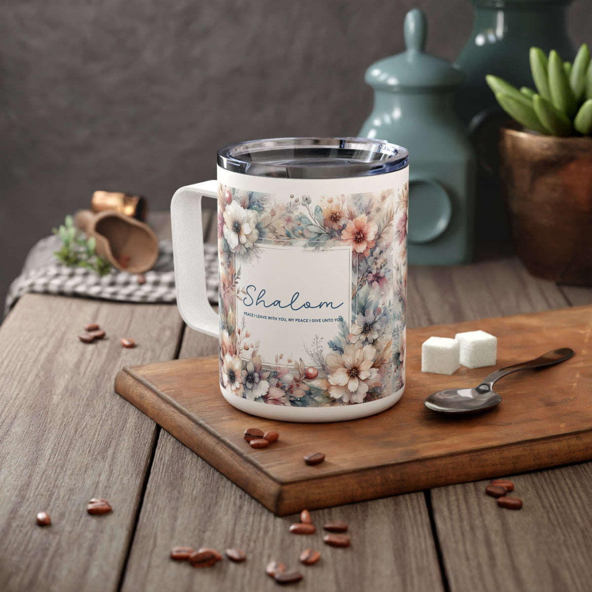 A white stainless steel coffee mug with a floral print and the word 'Shalom' on the front, styled on a wooden cutting board with coffee beans, sugar cubes, and a spoon on a rustic wooden table.