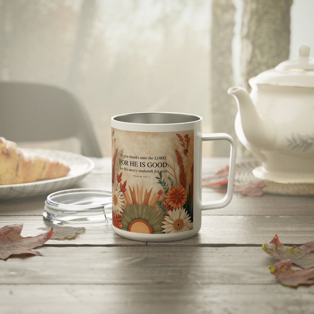 A white stainless steel mug with a floral and inspirational print sits on a rustic wooden table. The print features the text 'God is good even when feels bad' surrounded by autumn flowers and leaves.