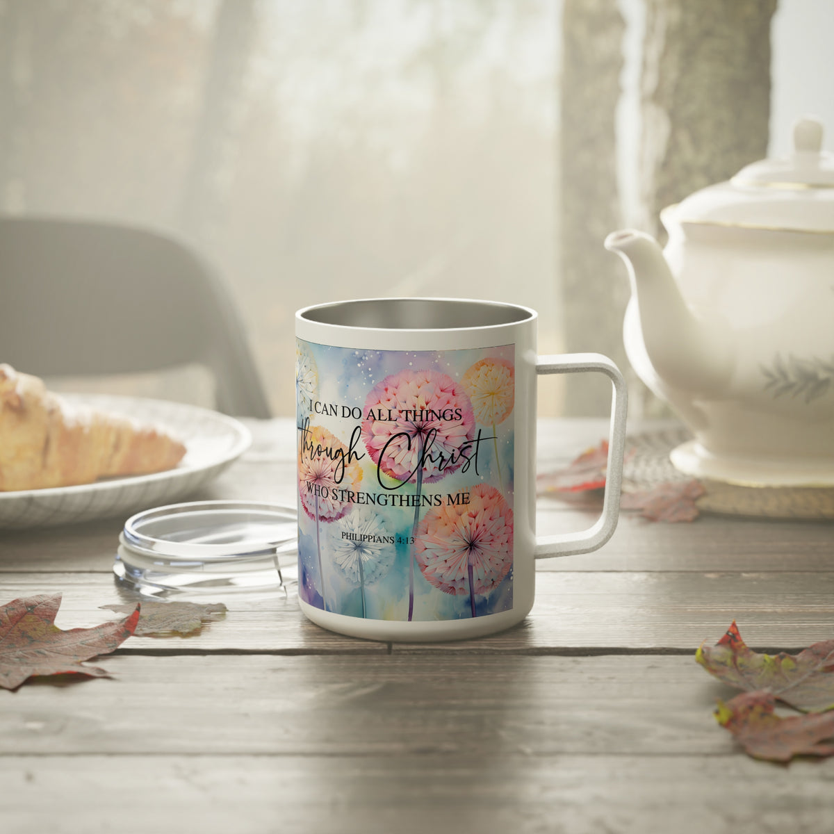 A white stainless steel  mug with a colorful dandelion illustration and the Bible verse Philippians 4:13 printed on it, sitting on a rustic wooden table with a glass lid, a teapot, and a pastry in the background.