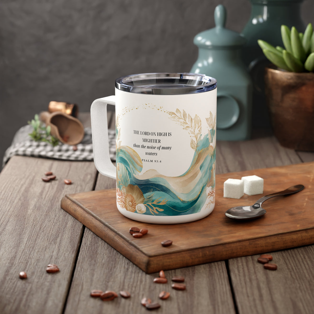 A white ceramic coffee mug with a gold and teal wave design and scripture text, displayed on a wooden cutting board with coffee beans, sugar cubes, and a spoon in a kitchen setting.
