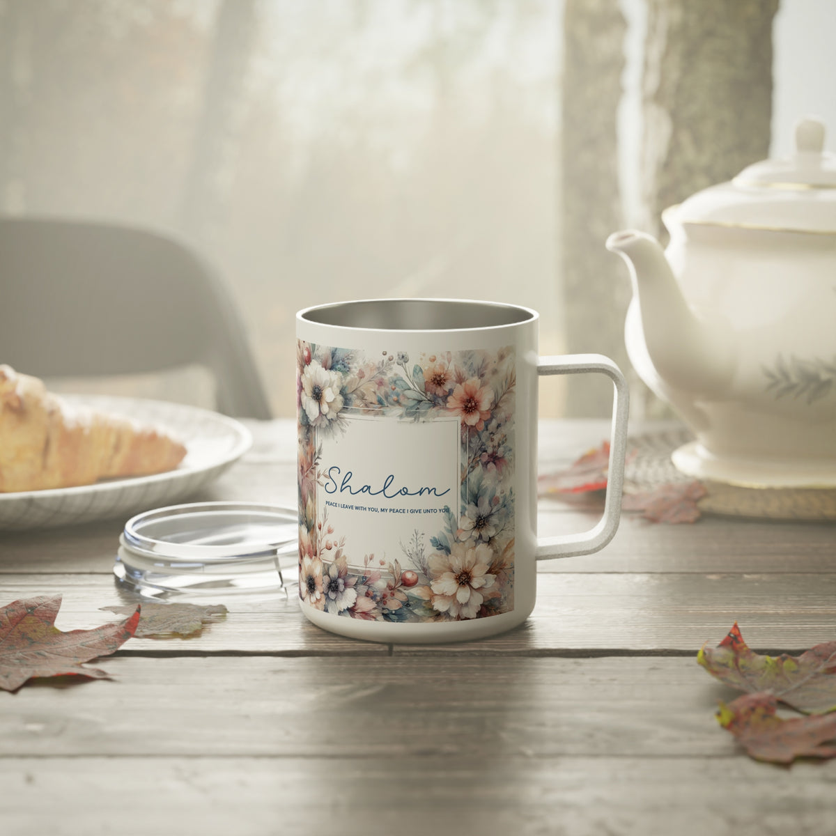 A stainless steel  mug with a floral border and the word 'Shalom' printed on the front, styled on a wooden table with a teapot, plate, and leaves in a softly lit interior.