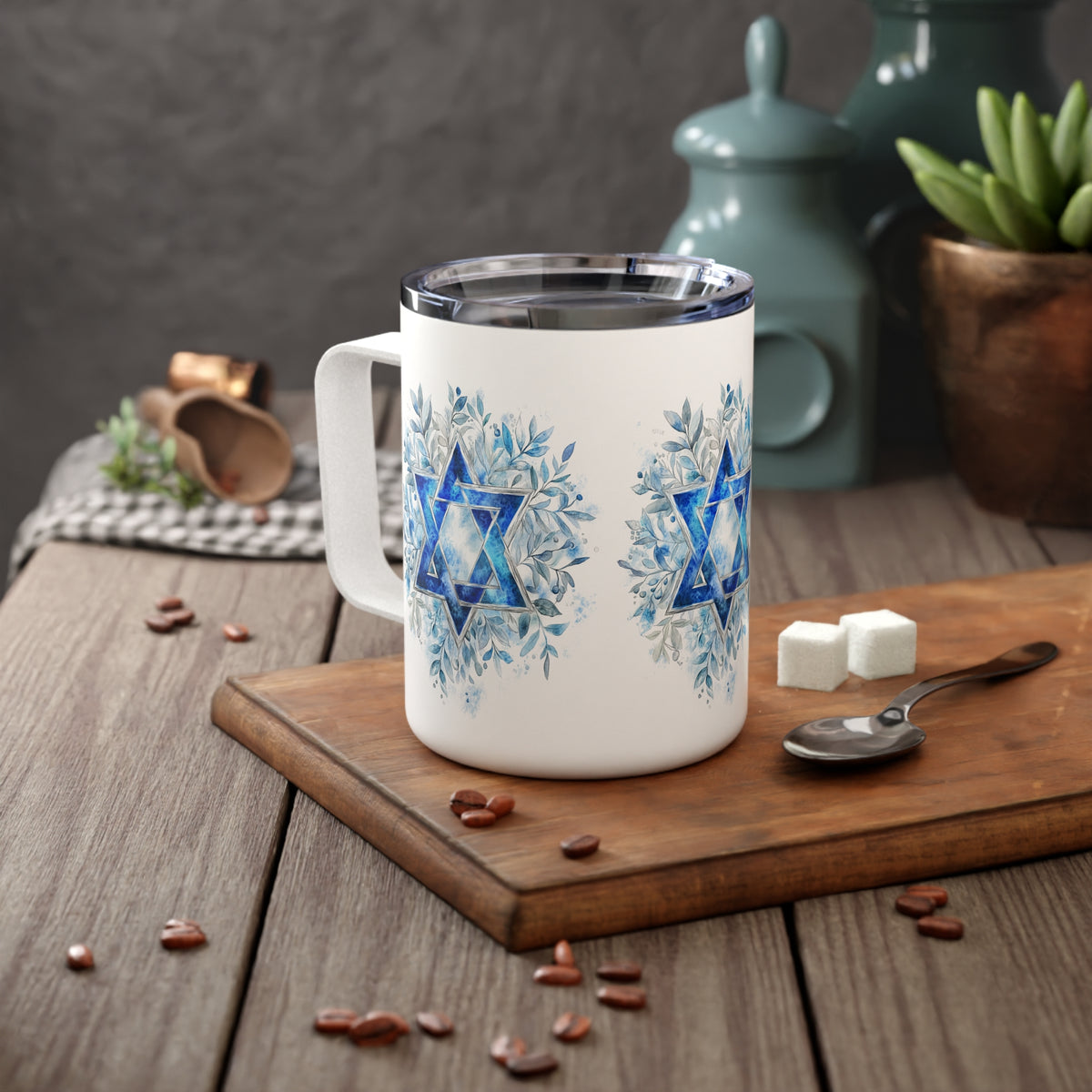 A white insulated mug with a blue Star of David and floral snowflake design, shown on a wooden cutting board with coffee beans, sugar cubes, and a spoon in a styled tabletop setting.