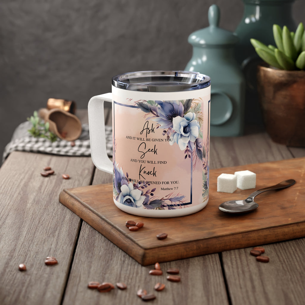 A white insulated coffee mug with a floral print and Bible verse, displayed on a wooden cutting board with coffee beans, sugar cubes, and a spoon in a kitchen setting.