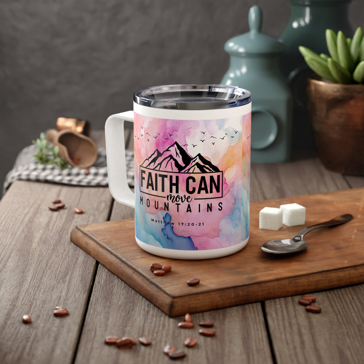 A white insulated mug with a colorful watercolor mountain design and the text 'FAITH CAN move MOUNTAINS' sits on a wooden cutting board on a rustic table, with coffee beans, sugar cubes, a spoon, and teal ceramic containers in the background.