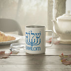 A white stainless steel  mug with a blue menorah graphic and the word 'SHALOM' printed on it, set on a wooden table in a cozy kitchen or cafe environment with a teapot, plate, and leaves nearby.