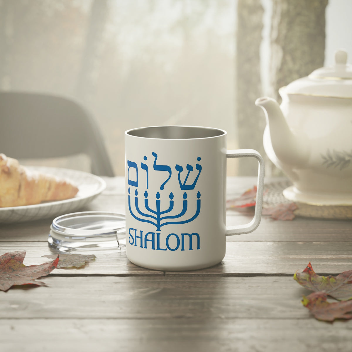 A white stainless steel  mug with a blue menorah graphic and the word 'SHALOM' printed on it, set on a wooden table in a cozy kitchen or cafe environment with a teapot, plate, and leaves nearby.