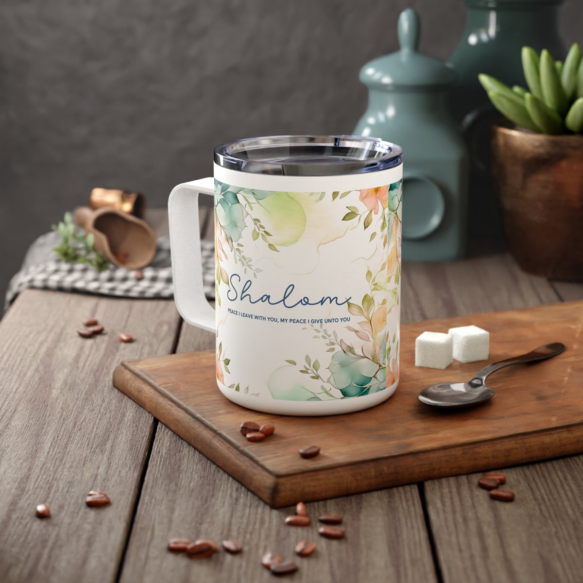 A white stainless steel insulated mug with a floral watercolor design and the word 'Shalom' printed on the side, styled on a wooden cutting board with coffee beans, sugar cubes, and a spoon in a kitchen setting.