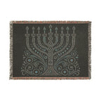 A rectangular woven throw blanket with fringe on all sides, featuring a large menorah and ornate scrollwork design in light gray and turquoise on a dark charcoal background.