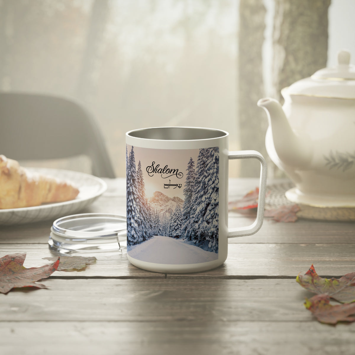 A white stainless steel mug with a custom printed winter forest design and the name 'Slaton' on the front, set on a rustic wooden table with a glass lid, a teapot, and autumn leaves in a cozy, softly lit environment.
