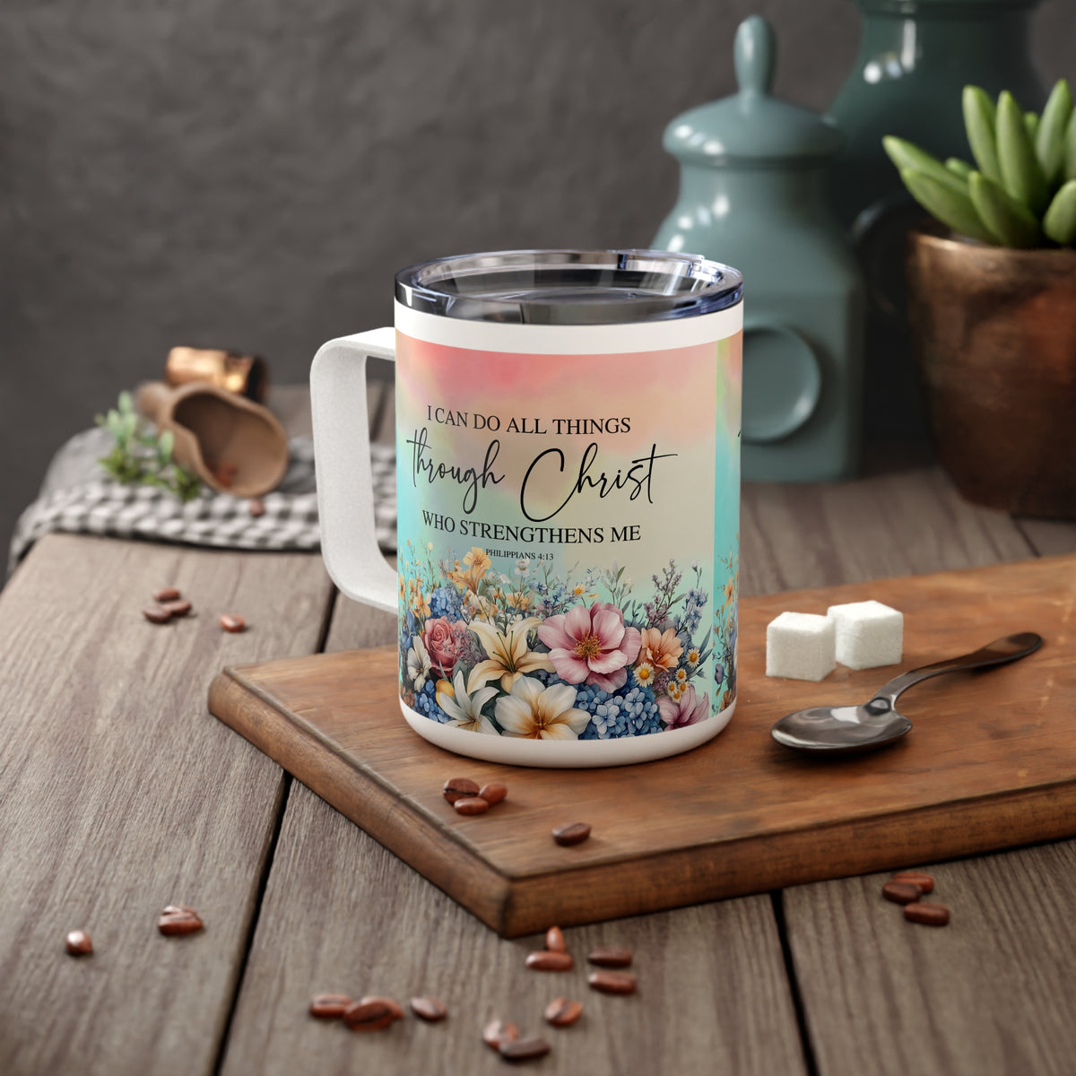A white insulated mug with a clear lid sits on a wooden cutting board on a rustic table. The mug features a pastel gradient background with the printed text 'I CAN DO ALL THINGS through Christ WHO STRENGTHENS ME' and a colorful floral illustration at the bottom.