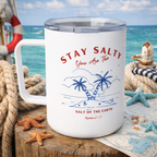 A white handled tumbler with a clear lid sits on a wooden dock with a beach and ocean backdrop. The tumbler is printed with the words 'STAY SALTY You Are The' and 'SALT OF THE EARTH' around a blue palm tree illustration.