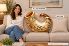 A woman sits on a beige sofa next to a large decorative shofar-shaped pillow. The pillow has an ornate gold, brown, and green leaf pattern.