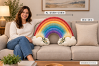 A woman sits on a beige sofa holding a large rainbow-shaped throw pillow with white cloud ends in a living room. White dimension lines label the pillow as XL 27.