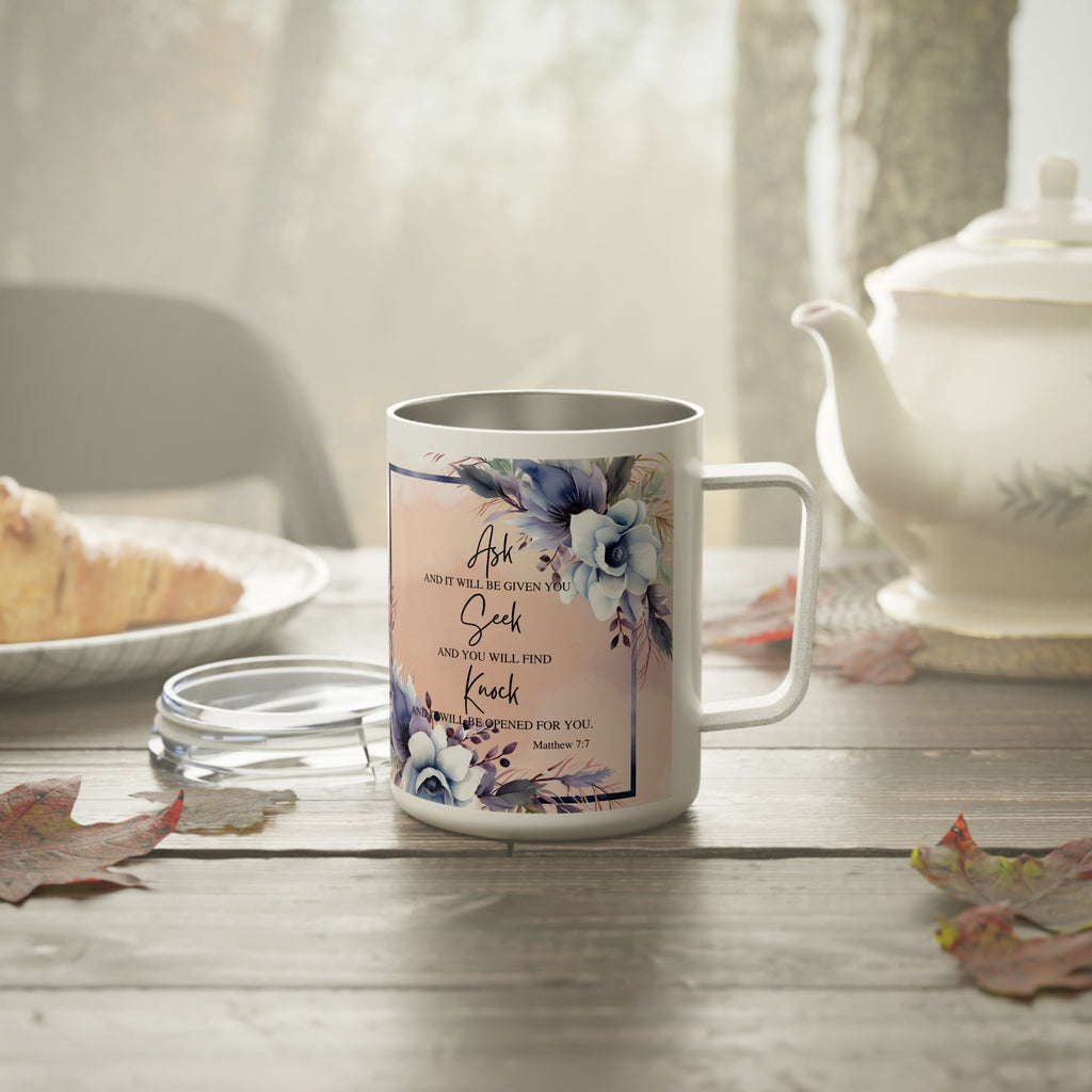 A white stainless steel coffee mug with a floral design and Bible verse print, set on a rustic wooden table next to a teapot and a glass lid.
