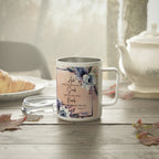 A white stainless steel coffee mug with a floral design and Bible verse print, set on a rustic wooden table next to a teapot and a glass lid.