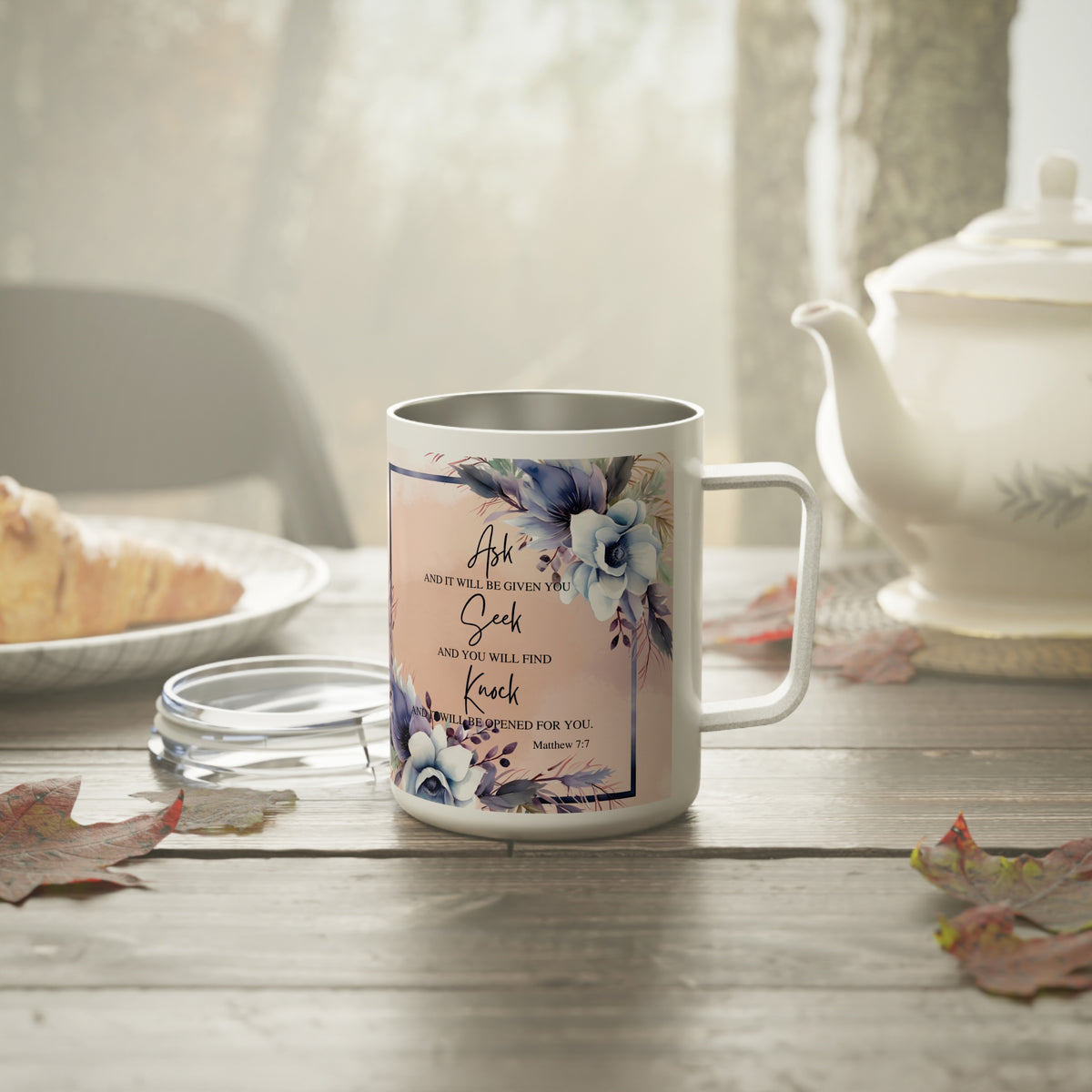 A white stainless steel coffee mug with a floral design and Bible verse print, set on a rustic wooden table next to a teapot and a glass lid.