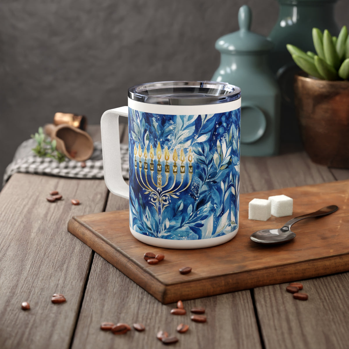 A white stainless steel mug with a blue floral pattern and a gold menorah design, placed on a wooden cutting board on a rustic table with coffee beans, sugar cubes, and a spoon.