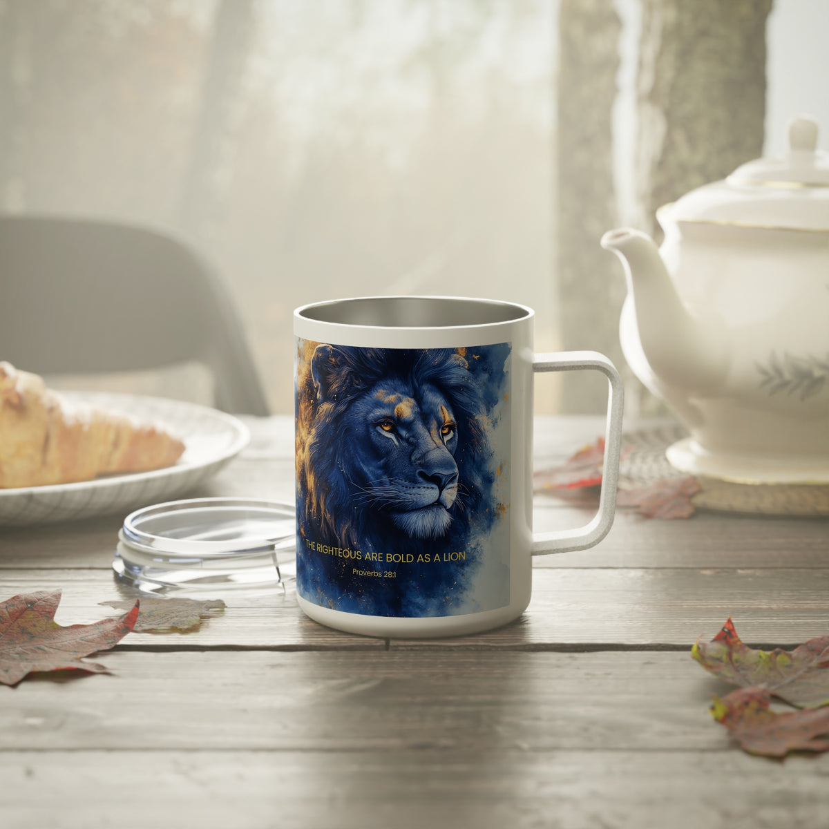 A white ceramic mug with a printed blue lion illustration and the quote 'THE RIGHTOUS ARE BOLD AS A LION Proverbs 16:42' sits on a wooden table in a cozy kitchen setting with a teapot, plate, and leaves.