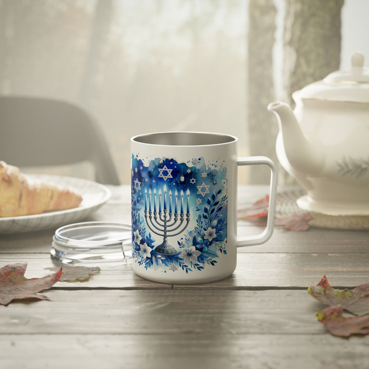 A white stainless steel mug with a blue Hanukkah menorah and floral design, set on a rustic wooden table with a teapot, plate, and leaves in a softly lit room.