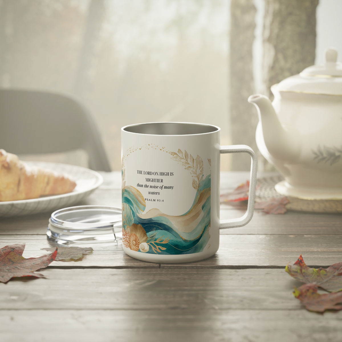 A white ceramic mug with a handle, featuring a printed design of teal and gold waves, a pearl, and a Bible verse, displayed on a wooden table with a teapot and plate in a softly lit kitchen setting.