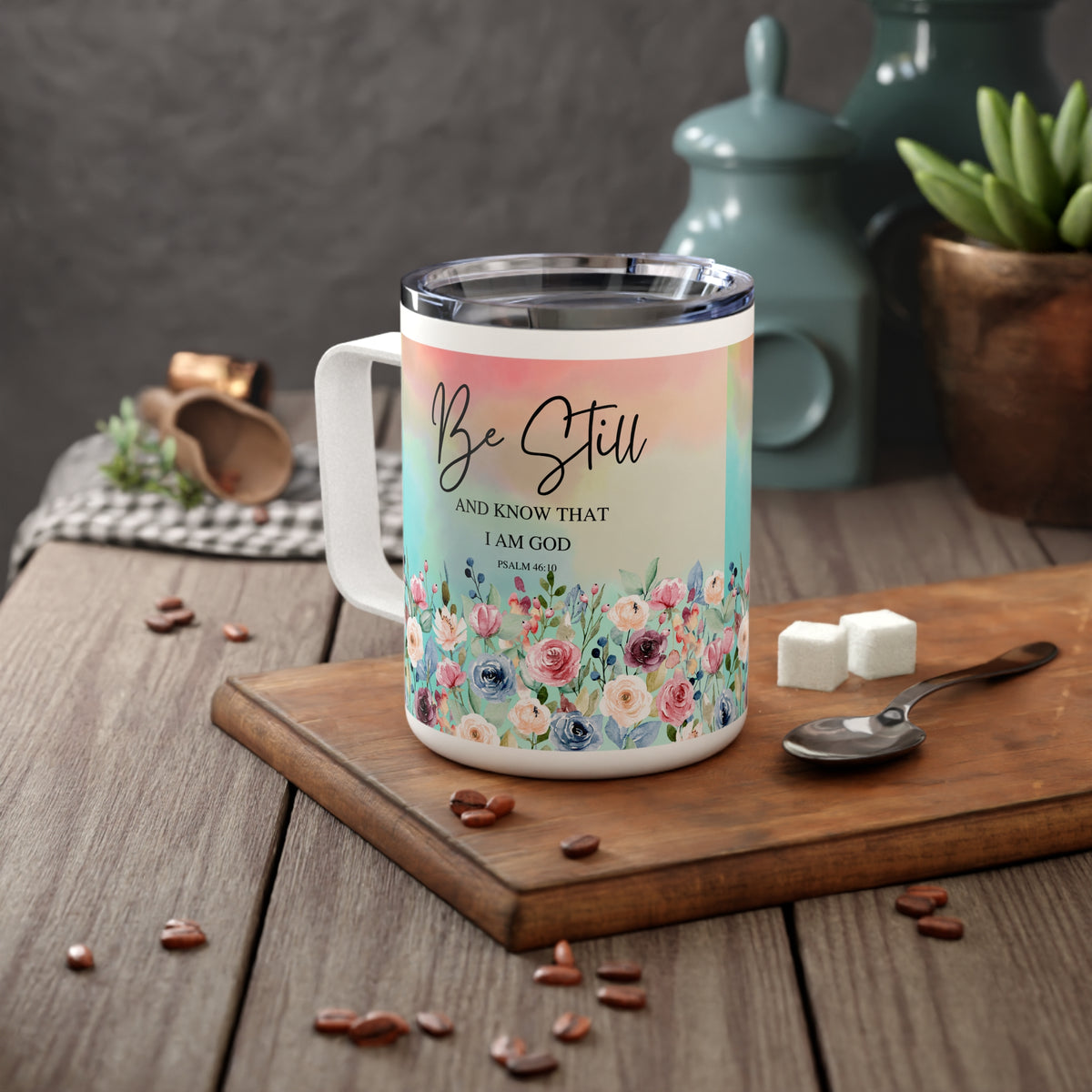 A white stainless steel travel mug with a colorful floral design and the text 'Be Still AND KNOW THAT I AM GOD' from Philippians 4:13, displayed on a wooden cutting board with coffee beans, sugar cubes, and a spoon in a kitchen setting.