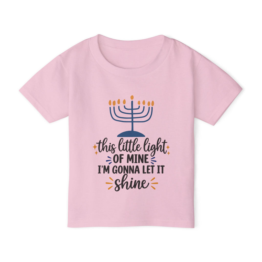 A light pink short-sleeve kids' t-shirt with a Hanukkah graphic on the front. The graphic features a blue menorah with orange flames and the text 'this little light OF MINE I'M GONNA LET IT shine' in black and orange script.