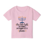 A light pink short-sleeve kids' t-shirt with a Hanukkah graphic on the front. The graphic features a blue menorah with orange flames and the text 'this little light OF MINE I'M GONNA LET IT shine' in black and orange script.