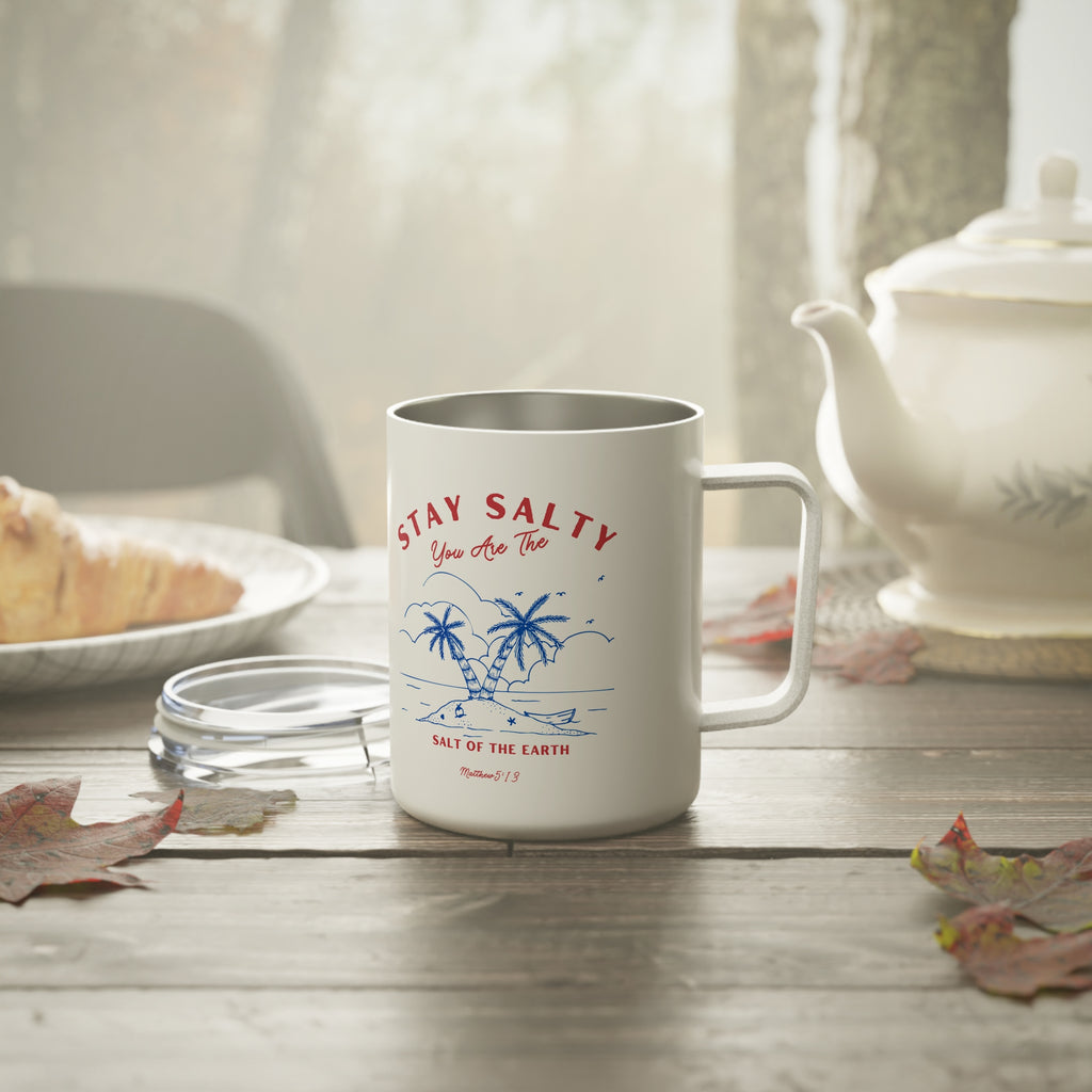 A white stainless steel coffee mug with a silver interior, featuring a blue and red tropical graphic with the text 'STAY SALTY You Are That' and 'SALT OF THE EARTH'. The mug is set on a rustic wooden table with a glass lid beside it, surrounded by autumn leaves, a plate of food, and a white teapot in the background.