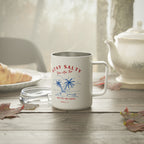 A white stainless steel coffee mug with a silver interior, featuring a blue and red tropical graphic with the text 'STAY SALTY You Are That' and 'SALT OF THE EARTH'. The mug is set on a rustic wooden table with a glass lid beside it, surrounded by autumn leaves, a plate of food, and a white teapot in the background.