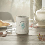 A white stainless steel  mug with a silver interior and handle, featuring a blue floral design and the text 'SHABAT VIBES ONLY' and 'MAKING TIME BY HEAVEN'S RHYTHM', set on a wooden table with a teapot and plate in a softly lit kitchen scene.