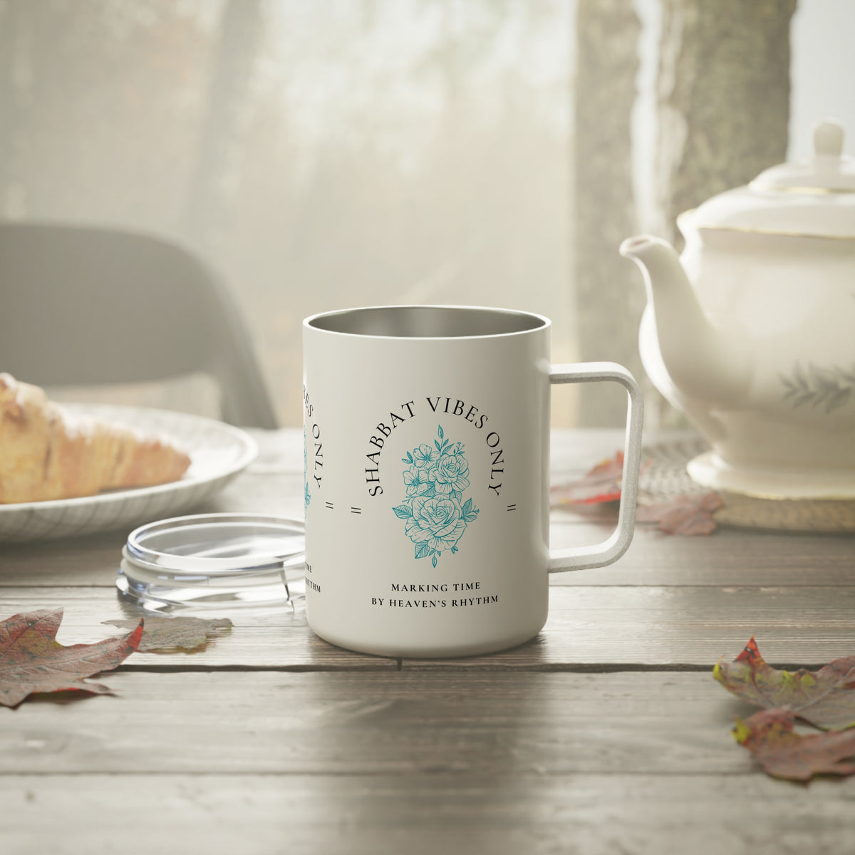 A white stainless steel  mug with a silver interior and handle, featuring a blue floral design and the text 'SHABAT VIBES ONLY' and 'MAKING TIME BY HEAVEN'S RHYTHM', set on a wooden table with a teapot and plate in a softly lit kitchen scene.